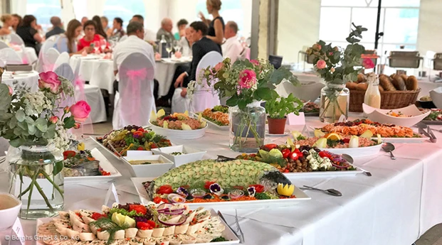 Buffet der Firma Borghs Catering & Event. Im Vordergrund ist ein kaltes Buffet aufgebaut. Im Hintergrund sitzen Menschen an stilvoll dekorierten Tischen in Bankettbestuhlung.