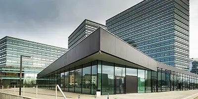 Gebäude in der seitlichen Ansicht. Im Vordergrund sieht man einen Vorplatz. Das Erdgeschoss des Gebäudes besteht aus zwei Glasfronten, die über Eck gehen. Im Hintergrund stehen Hochhäuser.