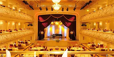 Festlicher Konzertsaal mit warmer Beleuchtung. In der Saalmitte stehen runde Banketttische, links und rechts erstrecken sich die Emporen über mehrere Stockwerke. Am Ende des Saals ist die Bühne, auf der ein Flügel steht. Die Bühne wird durch schwere, rote Vorhänge eingerahmt. Bild zur Verfügung gestellt von Kaisersaal, Erfurt