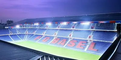 Das Stadion des FC Basel aus der Vogelperspektive. Man sieht die Tribüne mit dem Schriftzug FC Basel auf den unteren Rängen, der sich fast über die komplette Breite des Stadions zieht. Das Flutlicht ist angeschaltet und beleuchtet Spielfeld und Ränge. Bild zur Verfügung gestellt von Wassermann & Company AG Stadion, St. Jakob-Park
