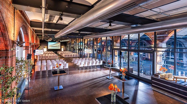 Location von Nord Event im Industrie-Stil mit roten Backsteinwänden und großer Fensterfront. Im Vordergrund stehen schwarze Stehtische mit orangenen Blumen. Im Hintergrund sind Stuhlreihen mit weißen Stühlen, in Richtung Podium mit Leinwand aufgestellt.