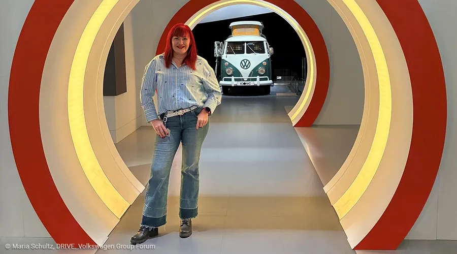 Eine Frau steht auf einem langen Gang, der durch eine Art Torbögen eingefasst ist. Der äußere Rand der Bögen ist rot eingefasst, in den Bögen ist ein Leuchtstreifen. Im Hintergrund ist ein alter VW-Bus zu sehen.
